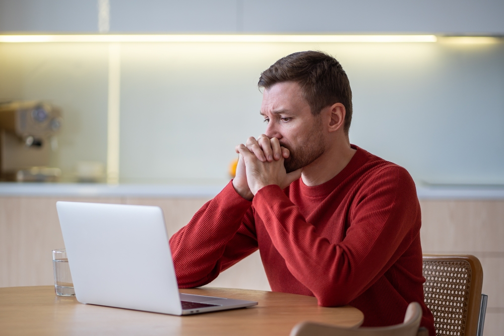 Sad frustrated man procrastinating after fired from job looking at laptop screen on kitchen at home
